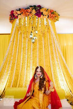 a woman sitting on a bed in front of a yellow curtain with flowers hanging from it