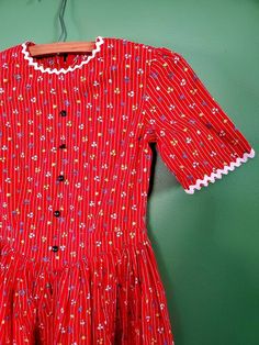 "Vintage 50s Girls Calico Cotton Dress w/ Ric Rac Trim. Super cute Red floral and striped dress with gray blue buttons (just for decoration - doesn't button) and white ric rac trim. The gathered skirt is very full and drops slightly in the front from the waist. The accent colors on fabric are yellow, white, blue and green. The dress is unlined with a back zipper. See condition below. II SPECIFICS II Brand: unknown Made in: unknown Materials: calico cotton  II MEASUREMENTS II Tag size: No tag so please see measurements below.  Length: 37\"  Waist: 25\"  Sleeve: 9\" Chest: 29\"   Feel free to contact me for any additional measurement.   II CONDITION II Great condition with no stains or holes.                                                                  Please keep in mind that there may Red Retro Vintage Dress For Spring, Spring Vintage Cotton Dress With Buttons, Retro Striped Dresses With Buttons, Striped Retro Dresses With Buttons, Cute Fitted Cotton Vintage Dress, Striped Fitted Dress With Buttons, Fitted Striped Dresses With Buttons, Retro Cotton Vintage Dress With Buttons, Retro Cotton Dress With Buttons