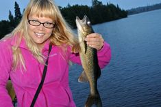 a woman in pink jacket holding up a small fish on a lake with trees in the background