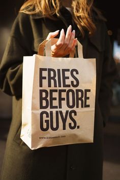 a woman holding a paper bag with the words fries before guys printed on it in black