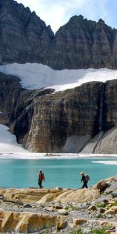 two people are walking on the rocks near a mountain lake and some snow covered mountains