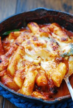 a skillet filled with pasta and sauce on top of a blue cloth next to a spoon