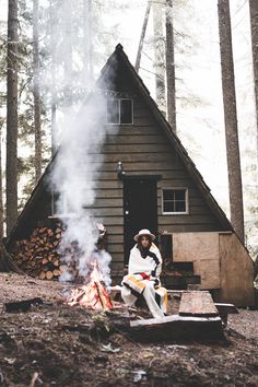 a man sitting next to a campfire in front of a small cabin surrounded by trees