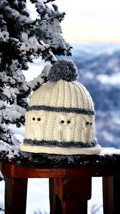 a white knitted hat sitting on top of a wooden table next to snow covered trees