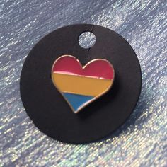 a heart shaped pin sitting on top of a black circle with a red, yellow and blue stripe