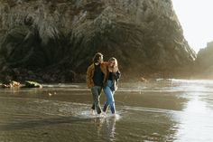 two people walking on the beach in front of a rock formation at low tide, with one person holding his arm around the other's shoulder