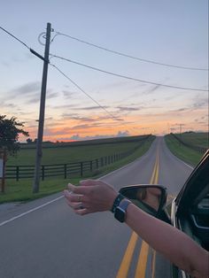 a person holding their hand out the window of a car on a country road at sunset