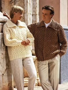 a man and woman standing next to each other near a stone wall wearing sweaters and pants