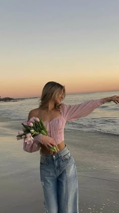 a woman is standing on the beach with flowers in her hand and pointing at the water