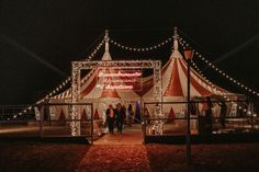 a circus tent is lit up at night with lights on the sides and people walking under it