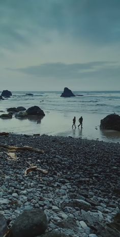 two people are walking on the beach with rocks and water in the foreground, while one person is holding an umbrella