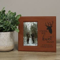 a wooden frame with an image of two men and a deer in the woods, on a table next to a potted plant