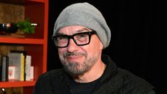 a man wearing glasses and a beanie looks at the camera while standing in front of a bookshelf