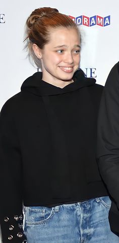 a young woman with red hair wearing a black hoodie and jeans, smiling at the camera