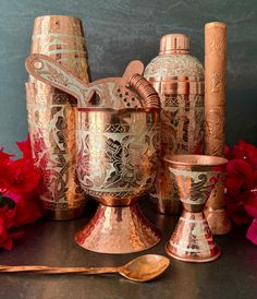 an assortment of copper colored vases and utensils with red flowers in the background