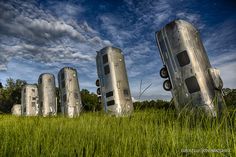 an image of some old cars in the middle of a grassy field with trees and clouds behind them