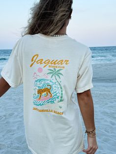 Our Sunhoney Jaguar Beach Club T-shirt is our newest exclusive design! This fun, funky design features bright pastel colors with a surfing Jaguar on the front and back catching a wave! This design is printed on Comfort Colors so you know it's going to be super comfy! Join the Jaguar Beach Club and rock this tee all summer long! Designed by us, handmade by us in Jacksonville Beach, FL Content: 100% Cotton Fit: TTS - size up for oversized fit Model wearing size: M Model sizing: 0-2 top, 24" bottom Funky Beach Outfits, Retro Summer Tops With Back Print, Surfing Top With Front Print For Beach Season, Graphic Tee With Screen Print For Vacation, Summer Graphic Tee With Back Print, Cotton Beach Top With Back Print, Cotton Top With Back Print For Beach, Graphic Tee Tops With Back Print For Beach, Summer Beach Tops With Back Print