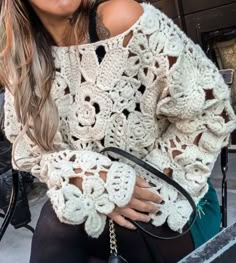 a woman sitting on a bench wearing a white crochet sweater and holding a black handbag