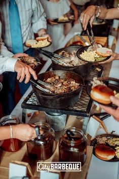 several people are serving themselves food from trays on a buffet table with hot dogs and hamburgers