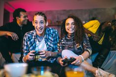 two people sitting on a couch playing video games with other people in the back ground