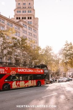 a red double decker bus driving down a street in front of tall buildings and trees