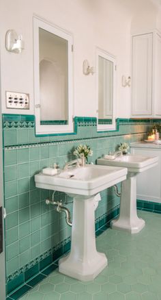 two white pedestal sinks in a green tiled bathroom with mirrors on the wall and lights above them