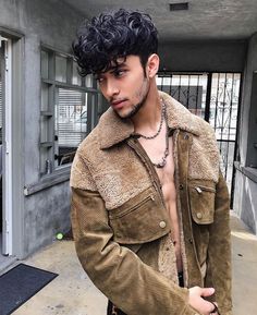 a man with curly hair and no shirt wearing a brown jacket standing in front of a building