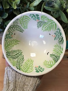 a hand holding a green and white bowl with leaves painted on the bottom, in front of a potted plant