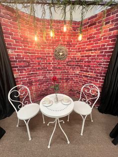 two white chairs and a table in front of a brick wall with lights on it