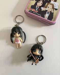 two anime keychains sitting next to each other on a white surface with a tin case in the background