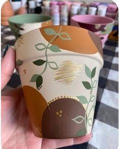 a hand holding up a paper cup with leaves and circles painted on the outside, along with other cups in the background