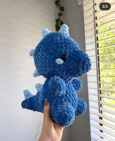 a hand holding up a blue crocheted stuffed animal in front of a window