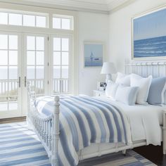 a white bed with blue and white striped comforter in a bedroom next to large windows