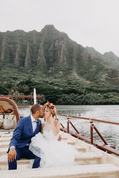 a bride and groom are sitting on steps near the water with mountains in the background