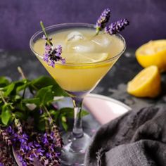 a drink in a martini glass with lavender sprigs and lemons on the side