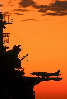 an airplane is flying in the sky over a ship at sunset or sunrise, with another plane on the deck