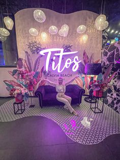 a woman sitting on a purple couch in front of a neon sign that reads tios