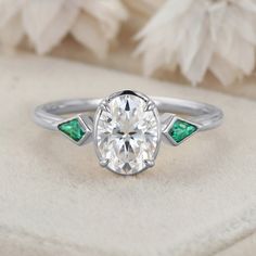 a diamond and emerald engagement ring sitting on top of a white cloth with feathers in the background