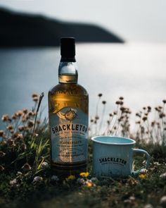 a bottle of shackleton next to a mug on the grass near water with wildflowers