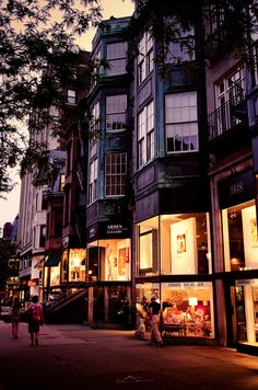 people are walking down the street in front of some tall buildings at night with lights on