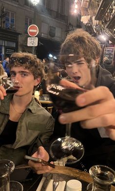 two young men sitting at a table with wine glasses in their hands and one holding a cell phone up to his mouth