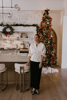 a woman standing in front of a kitchen counter next to a christmas tree with lights on it