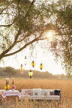 a picnic table set up under a tree in the middle of a field with lanterns hanging from it