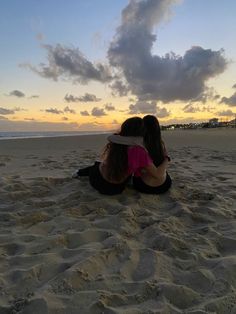 two women sitting in the sand at sunset with their arms around each other and looking into the distance