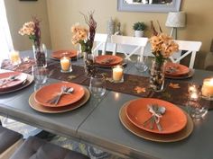 a dining room table set with orange plates and place settings, candles and flowers in vases