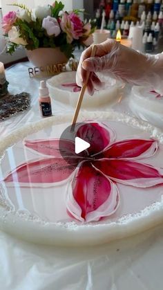 a person painting a flower on a white plate with pink flowers in the background and candles