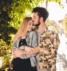 a man and woman standing next to each other in front of a tree with their arms around each other