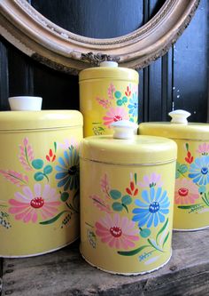 three yellow canisters with flowers painted on them sitting next to a round mirror