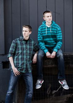 two young men sitting next to each other in front of a black wall and fence