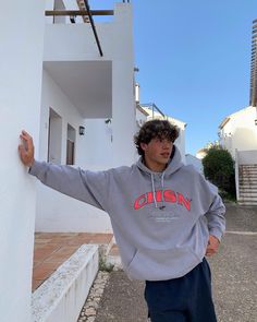 a young man leaning against the side of a white building wearing a grey hoodie